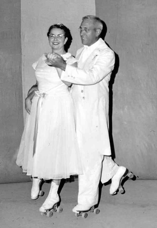 Two people in white outfits roller skate indoors, smiling and holding hands.